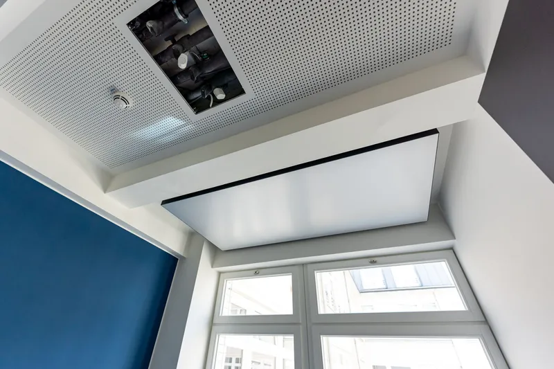 Detail der weißen Akustikdecke in der Kaisergalerie Hamburg mit geöffneter Revisionsklappe über der Hydraulik, abgehängtem interpanel Klimapanel und blauer Akzentwand am Fenster.