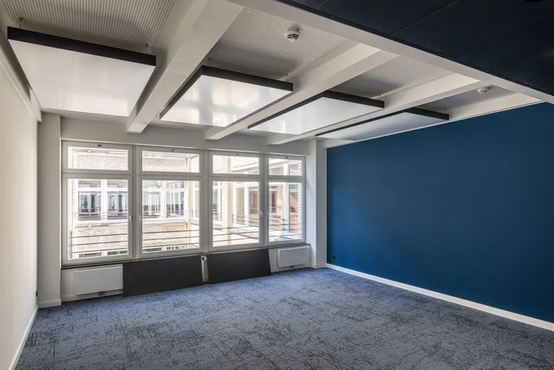 Büroraum der Kaisergalerie Hamburg mit blauer Akzentwand, abgehängten interpanel Klimapanels zwischen weißen Trägern und Eckfenstern zum Innenhof.
