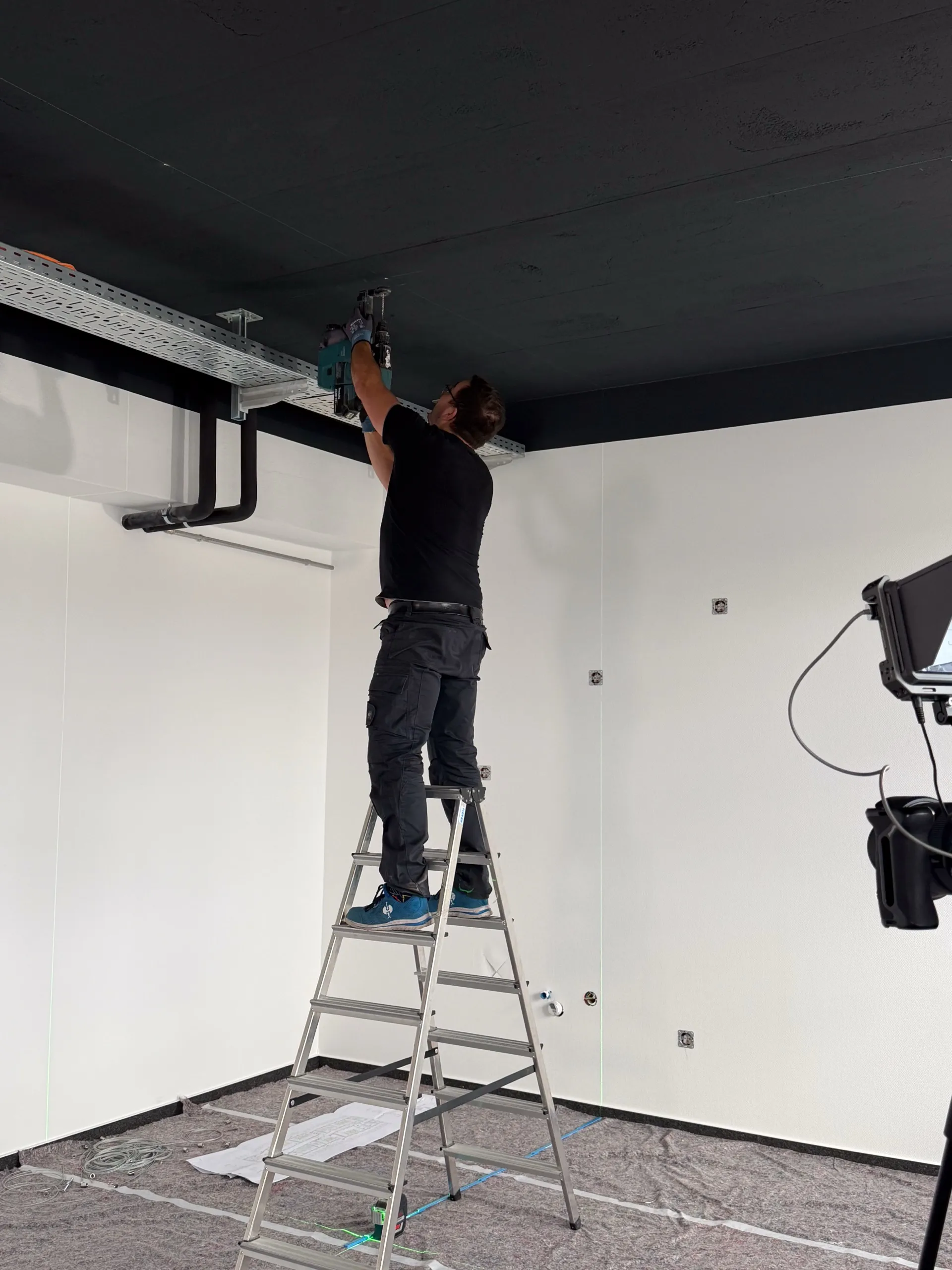 Monteur befestigt auf einer Leiter den Deckenanker für ein interpanel Deckensegel an der Rohdecke