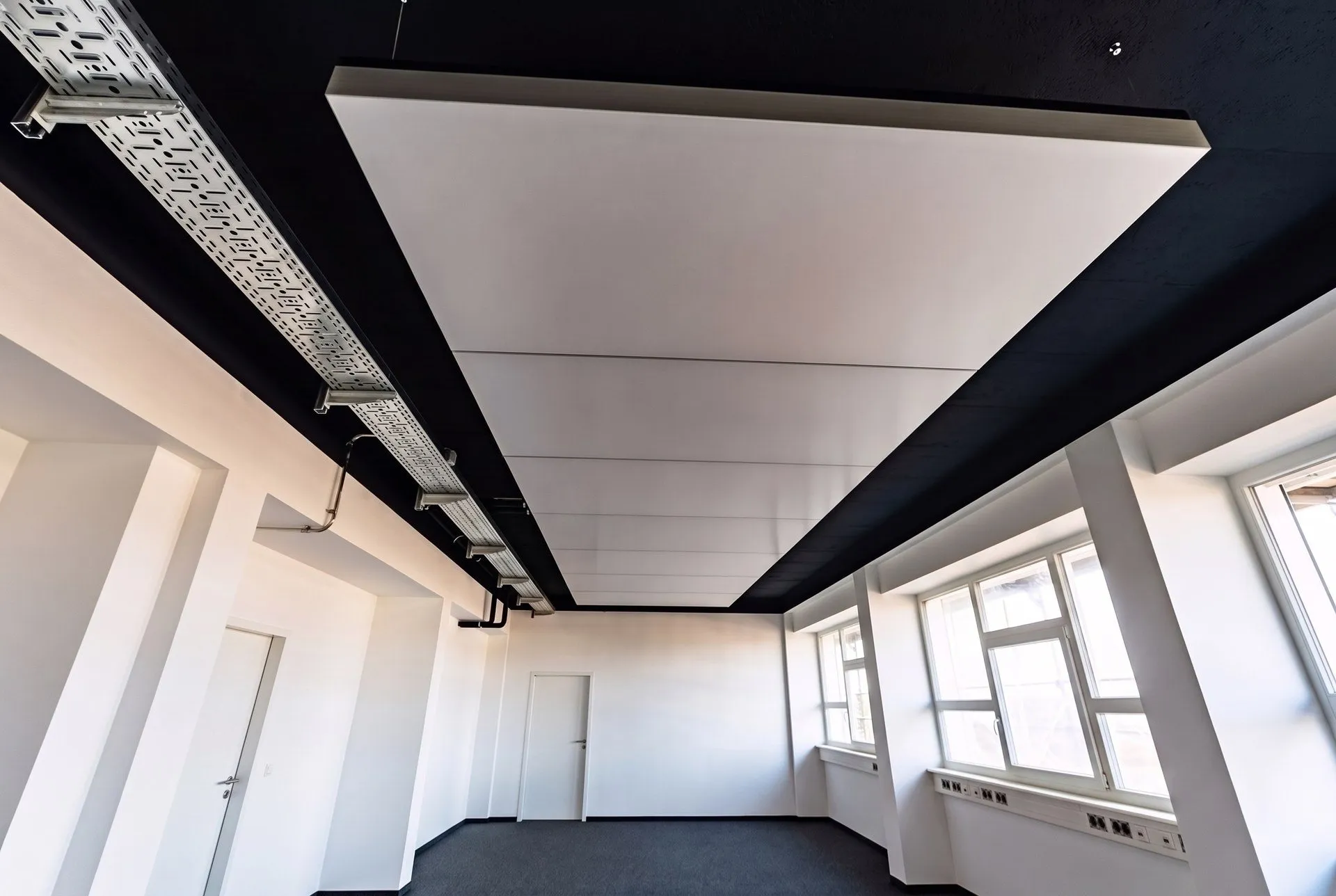 Abgependeltes interpanel Deckensegel mit Aluminiumrahmen unter der schwarzen Rohdecke, daneben Kabeltrassen