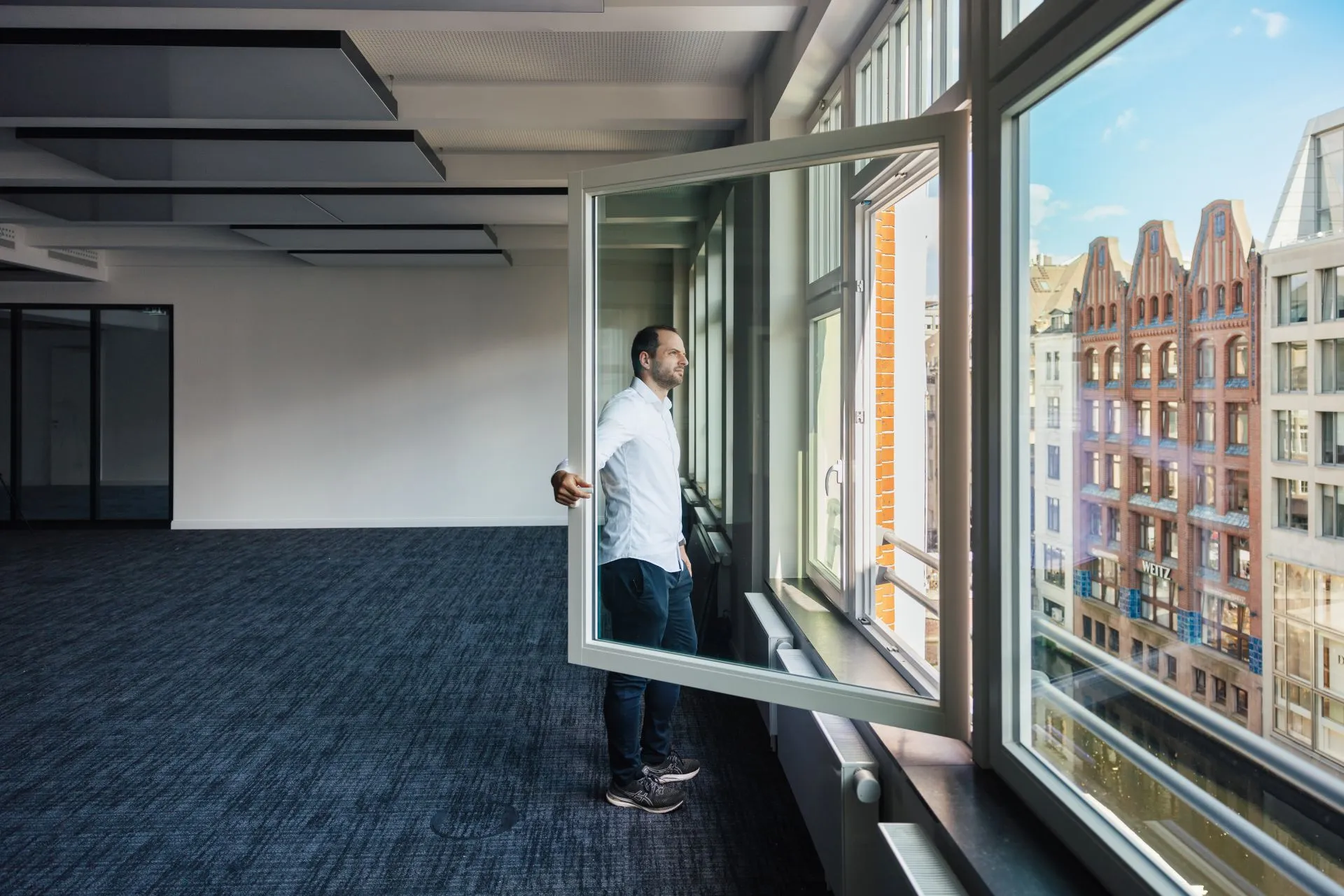 Mitarbeiter öffnet das Fenster in einem Bestandsbüro mit interpanel Deckensegeln, Blick auf die Hamburger Speicherstadt