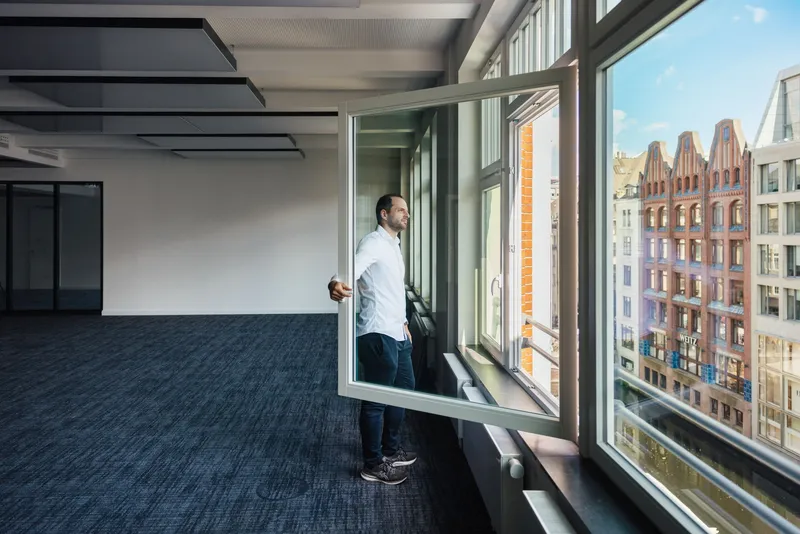 Person am geöffneten Fenster der Kaisergalerie Hamburg mit Blick auf Hamburger Backsteinfassaden, abgehängten interpanel Klimapanels und blauem Teppichboden.